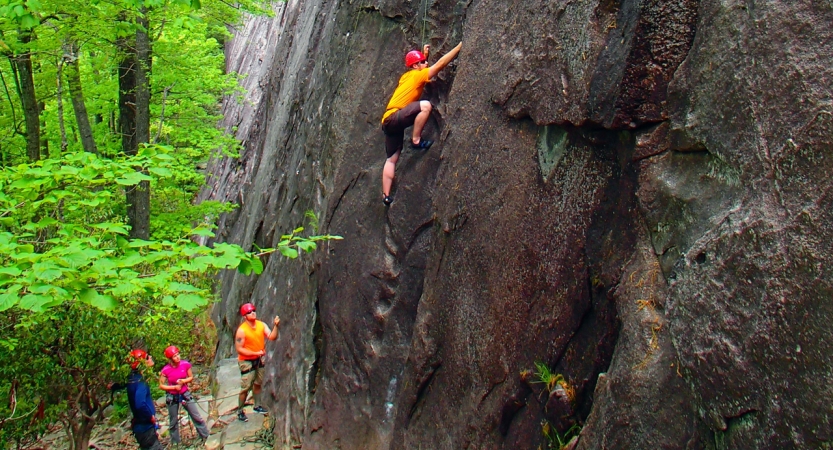 A person wearing safety gear is secured by ropes as they climb a rock wall. On the ground, one person belays while others look on. 