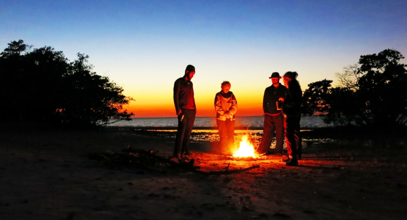 Four people stand around a campfire on a beach. The light appears in shades of blue, yellow and orange, as if the sun is setting or rising.