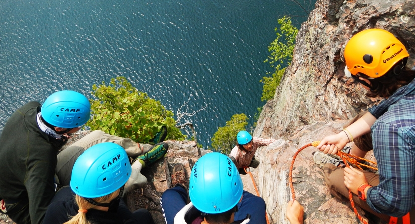 A group of students wearing helmets look over the edge of a cliff at a rock climber below. There is blue water below them.