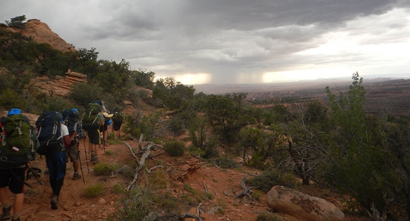 A group of people hike along a trail in a desert landscape. In the distance, it appears to be raining. 