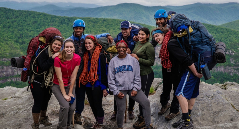 Group backpacking tripsA group of students stand on a rock overlook and smile at the camera. There is a green mountainous landscape behind them.