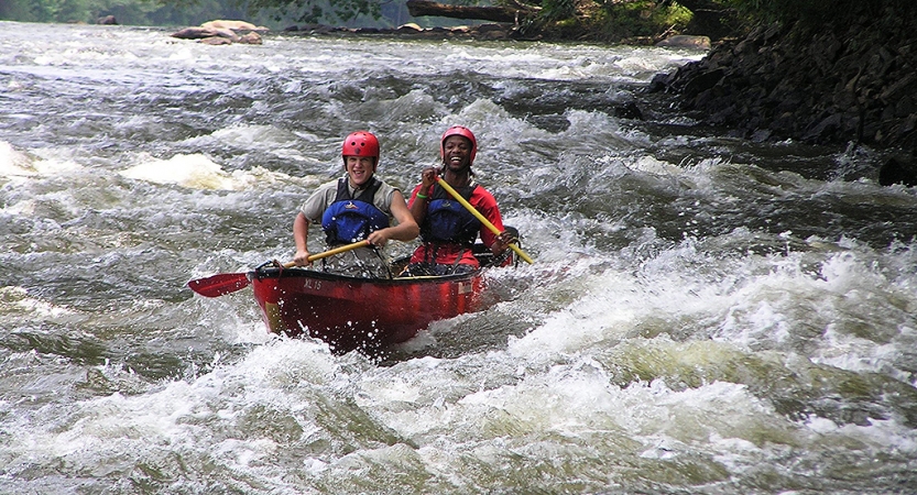 two students wearing safety gear navigate a canoe through whitewater in north carolina on an outward bound trip