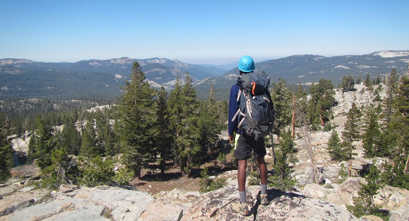 a person wearing a helmet and a backpack faces away from the camera and looks out over a vast mountainous landscape