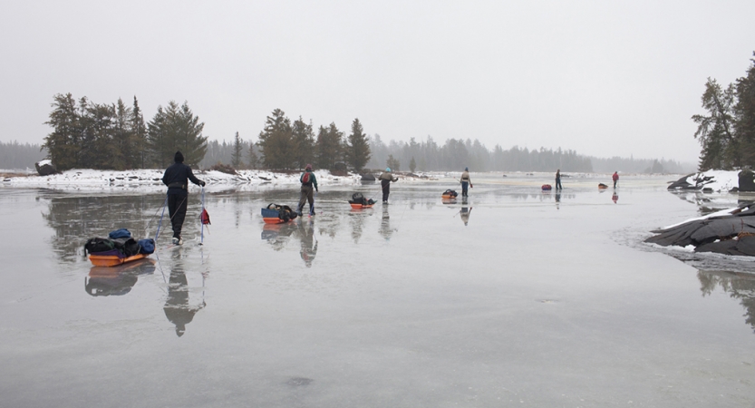 A line of people pull small sleds in a line across an icy lake