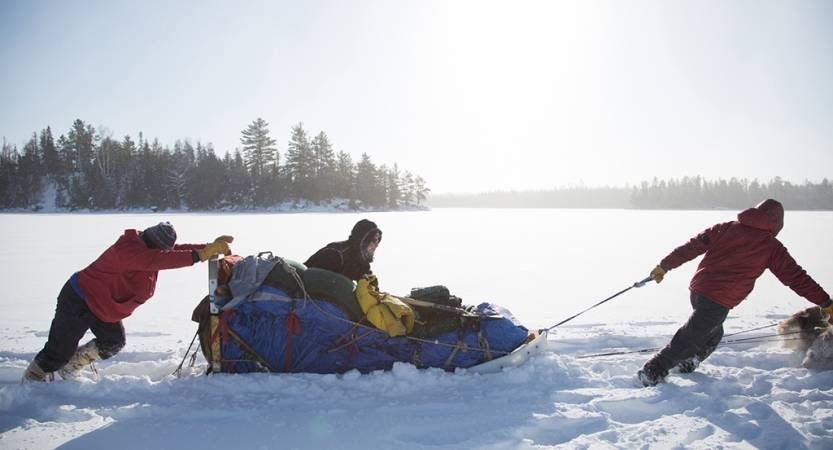 Three mushers help to push and pull a sled through thick snow. There are trees in the background. 